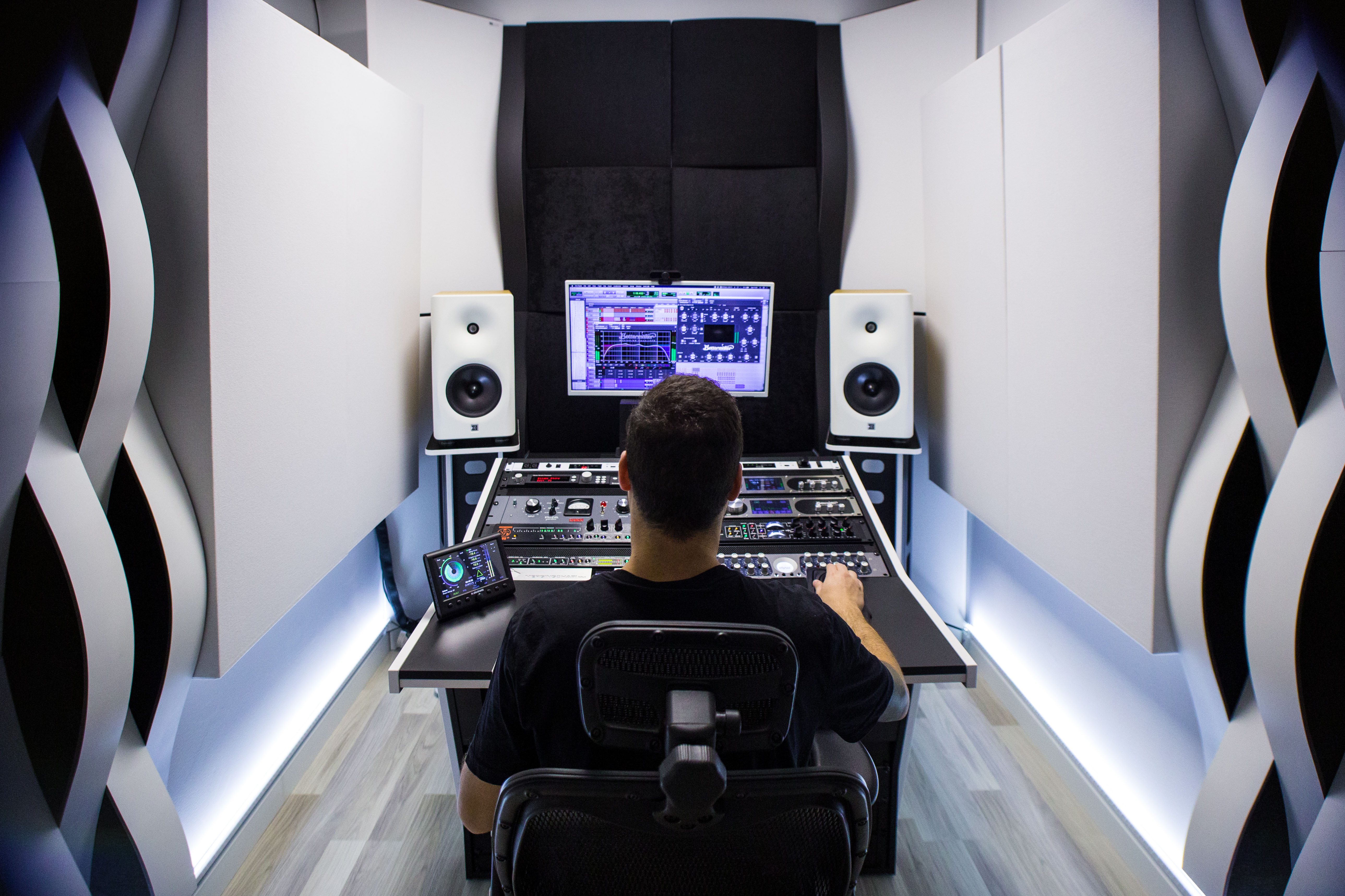 Salva Ballesteros seated on a studio chair at the mixing desk, facing two white monitor speakers and a computer screen with audio software open, surrounded by white and black acoustic wall panels in his mastering studio.
